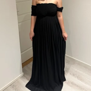 Svart axelbandslös balklänning  - Elegant svart axelbandslös långklänning med veckad design. Klänningen har en smickrande passform och faller vackert längs kroppen.