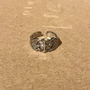 Elysian Fleur Cross Ring - En vacker ring med ett korsmotiv i mitten, omgiven av detaljrika bladmotiv som ger den en elegant och stilfull karaktär. Storlek US 8, men delvis justerbar för en flexibel passform. En ring som kombinerar både symbolik och estetik på ett harmoniskt sätt, perfekt för den som söker något både unikt och tidlöst