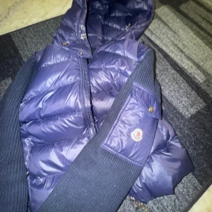 Blå cardigan från Moncler - Säljer en snygg blå dunväst från Moncler med dragkedja och huva. Västen har ribbade detaljer och en klassisk design som passar perfekt för kyligare dagar. Perfekt för dig som vill ha både stil och funktion.
