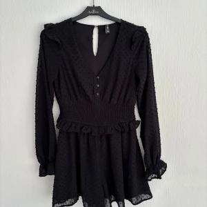 Svart klänning  - Snygg svart klänning från FB Sister med prickigt mönster och volangdetaljer. Klänningen har en v-ringad design med knappar framtill och långa ärmar. Perfekt för en stilren look!