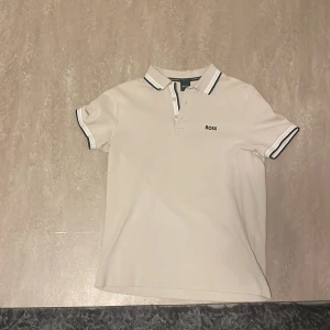 Beige kortärmad pikétröja från Boss - Säljer en beige pikétröja från Boss med klassisk krage och knappar. Tröjan har svarta och gröna detaljer på kragen och ärmsluten. Perfekt för en avslappnad men ändå snygg look.