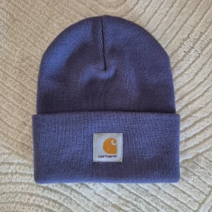 Lila mössa från Carhartt - Oanvänd mössa från Carhartt med uppvikt kant och logotyp framtill. Perfekt för kyliga dagar och en stilren look.