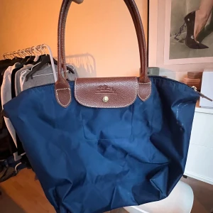 Blå shoppingväska med bruna detaljer - Snygg blå shoppingväska med bruna skinnhandtag och detaljer. Väskan har en praktisk dragkedja och en knapp för extra säkerhet. Perfekt för att bära med sig allt man behöver under dagen. Liknade longchmap 
