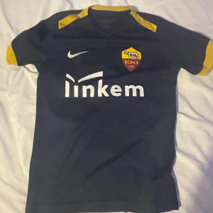 Svart och gul AS Roma fotbollströja - Säljer en svart AS Roma fotbollströja från Nike med gula detaljer på ärmar och krage. Tröjan har korta ärmar och klubbens emblem på bröstet. Perfekt för fotbollsfans!