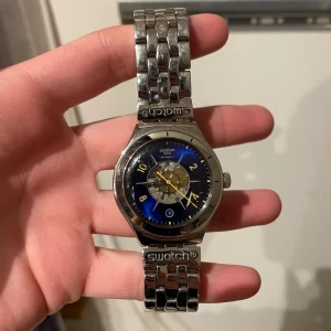 Blå och silver klocka från Swatch - Säljer en stilren klocka från Swatch med en blå urtavla och silverfärgad länkarmband. Klockan har en transparent baksida som visar urverket. Perfekt för den som gillar en kombination av klassisk och modern design. Ordinarie pris 1000kr