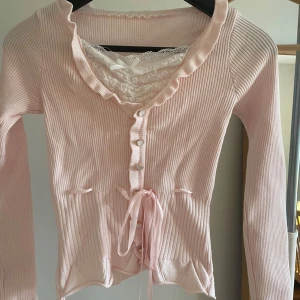 Rosa ribbad kofta med spetsdetaljer - Söt rosa ribbad kofta med knappar och knytband framtill. Har en vacker spetsdetalj vid halsringningen och volangkant. Perfekt för en romantisk look.