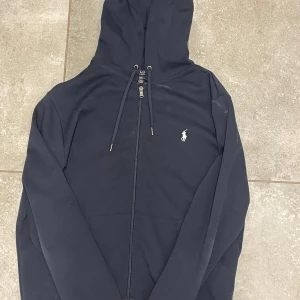 Svart hoodie från Polo Ralph Lauren - Säljer en svart hoodie från Polo Ralph Lauren med dragkedja och justerbar huva. Den har en liten broderad logga på bröstet. Perfekt för en avslappnad stil.