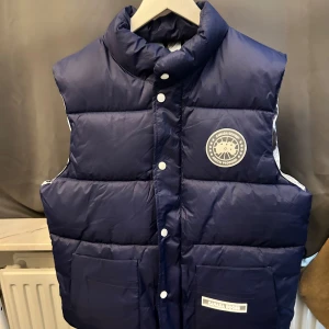 Blå dunväst från Canada Goose - Säljer en blå dunväst från Canada Goose med knappar och broderad logotyp på bröstet. Västen har en hög krage och två stora fickor framtill. Perfekt för kyliga dagar.