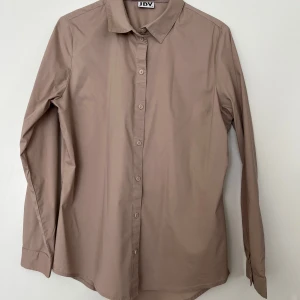 JDY ljusbrun skjorta 40 - Ljusbrun skjorta med långa ärmar och knäppning framtill, från JDY i storlek 40. Klassisk krage och rak passform. Skjortan är i nyskick. 72% bomull, 24% polyester och 4% elastan.   Mått: Längd ca 75 cm Byst ca 50 cm Midja ca 50 cm Ärmlängd mätt från axeln ca 60 cm Axelmått 40 cm
