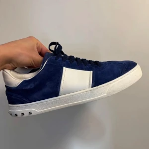 Blå och vita sneakers från Valentino - Snygga blå sneakers i mocka från Valentino med vita detaljer och snörning. Skorna har en rund tå och dekorativa nitar på hälen. Perfekta för en stilren look.
