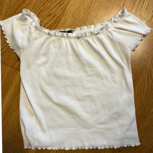 Vit ribbad offshoulder topp - Säljer en söt vit ribbad offshoulder topp med korta ärmar och volangdetaljer vid halsen på grund av att den inte kommer till användning. Den är använd men är i bra skick. Skriv om ni har frågor💕