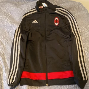Svart träningströja från Adidas - Säljer en svart träningsjacka från Adidas med AC Milan-logga på bröstet. Jackan har långa ärmar med klassiska gråa ränder längs sidorna och en dragkedja framtill. Den har också en röd detalj vid nederkanten. Perfekt för fotbollsfans!