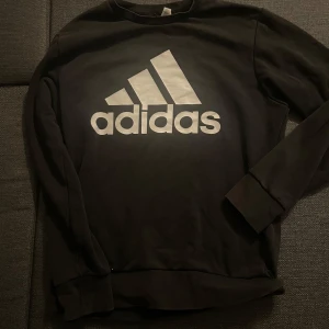 Svart tröja från Adidas - Säljer en svart tröja från Adidas med deras ikoniska logga i vitt på framsidan. Tröjan har långa ärmar och en rund halsringning. Perfekt för en sportig och avslappnad stil.