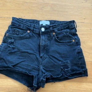 Svarta jeansshorts från H&M - Säljer ett par svarta jeansshorts från H&M med slitna detaljer och hög midja. Shortsen har en klassisk femficksdesign och knappgylf. Perfekta för en avslappnad stil.