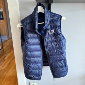 EA7 Väst (EMPORIO ARMANI) - En väldigt fin EA7 väst i marinblå. den är i väldigt bra skick och passar fint och bra in med allt ungefär. Det finns 2 fickor inuti västen. Jag är 181-182 ungefär och man ser på bilderna hur den sitter på mig. Den kommer inte med sin luva då den är köpt av en vän. Kom med frågor om det är något!