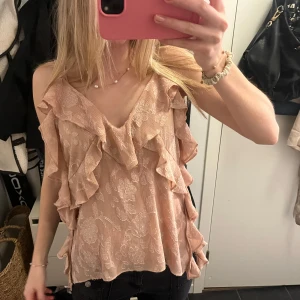 Beige blus med volanger - Säljer en söt beige blus med volanger och blommigt mönster. Blusen har en v-ringad design och är ärmlös, perfekt för varmare dagar. Passar bra till både jeans och kjol.