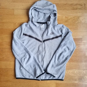 Grå huvtröja från Nike - Säljer en grå huvtröja från Nike med dragkedja och huva. Tröjan har ett svart och brunt randigt mönster över bröstet och Nike-loggan på vänster sida. Perfekt för en avslappnad stil. Pris kan diskuteras