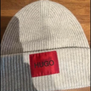 Grå ribbad mössa från Hugo Boss  - Snygg grå ribbad mössa från Hugo Boss med en röd etikett framtill. Perfekt för att hålla värmen under kyliga dagar.