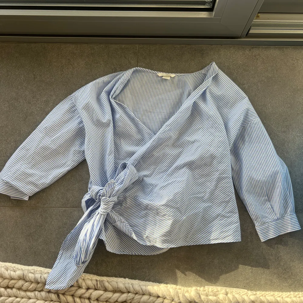 Säljer en snygg blå- och vitrandig omlottopp från H&M. Toppen har trekvartsärmar och ett knytband i midjan 💙. Yläosat.