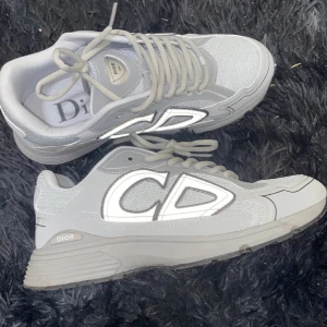 Grå sneakers från Dior - Snygga grå sneakers från Dior med stora CD-logotyper på sidorna. Skorna har en modern design med snörning och en bekväm sula. Perfekta för en stilren look.