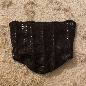 Svart paljettprydd tubtopp från Gina Tricot - Snygg svart tubtopp från Gina Tricot med glittrande paljetter och snörning i ryggen. Perfekt för en kväll ute eller festliga tillfällen. Toppen är axelbandslös och har en tight passform för en stilren look.