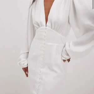 Vit långärmad klänning med knappar - Elegant vit klänning med v-ringning och långa ärmar. Klänningen har dekorativa knappar framtill och en dragkedja längs sidan. Aldrig använd, bara testad