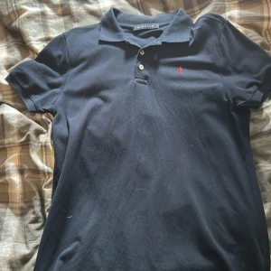 Svart pikétröja från Polo Club - Säljer en klassisk svart pikétröja från Polo Club med korta ärmar och en liten röd logga på bröstet. Perfekt för en stilren och avslappnad look. Tröjan har en krage och knappar framtill.