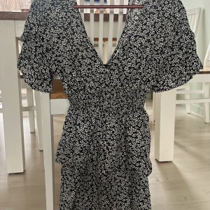 Svart blommig klänning - Säljer en svart klänning med vita blommor och volangdetaljer. Klänningen har en v-ringad design och korta ärmar. Perfekt för sommarens alla tillfällen!