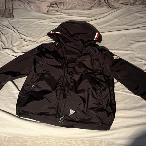 Svart vindjacka från Moncler - Säljer en stilren svart vindjacka från Moncler med huva och dragkedja. Jackan har Monclers logotyp på ärmen och detaljer i rött och vitt på huvan. Perfekt för blåsiga dagar och en snygg touch till din outfit. Jackan är i bra skick. 
