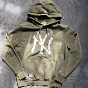 Olivgrön hoodie med New York Yankees-logga - Säljer en olivgrön hoodie från New Era med stor vit New York Yankees-logga på framsidan. Hoodien har en klassisk känguruficka och justerbar huva med snören. Storlek Xs men passar S. På bilden är jag 178 cm lång  och den sitter lite litet. Hoodien har en defekt som syns på tredje bilden. Vid frågor och funderingar är den bara att skicka dm💫