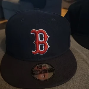 Svart keps från New Era - Snygg svart keps från New Era med ett broderat rött 'B' på framsidan. Kepsen är en 59FIFTY-modell och har en platt skärm. Perfekt för att ge din outfit en sportig touch.