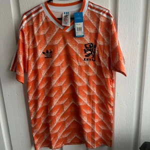 Orange fotbollströja från Adidas - Säljer en retroinspirerad orange fotbollströja från Adidas med ett unikt geometriskt mönster. Tröjan har korta ärmar och klassiska vita ränder på axlarna. KNVB-loggan och Adidas-märket pryder framsidan. Perfekt för fotbollsfans och samlare!