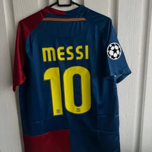 Blå och röd fotbollströja från Nike - Säljer en ikonisk fotbollströja från Nike i blått och rött med Messi och nummer 10 på ryggen. Tröjan har korta ärmar och ett tryck av Champions League-loggan på ärmen. Perfekt för fotbollsfans!