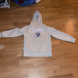 Beige hoodie från NBA - Säljer en beige hoodie från NBA med Los Angeles Lakers-logga på framsidan. Tröjan har en stor ficka framtill och långa ärmar. Perfekt för alla basketfans! 🏀