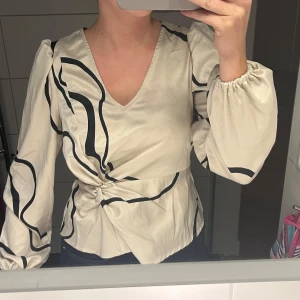 Beige omlottblus med svart mönster - Snygg beige omlottblus med svart abstrakt mönster. Blusen har långa ärmar med resår vid ärmsluten och en v-ringad hals. Perfekt för en stilren look.
