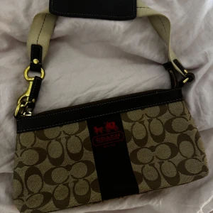 Beige och brun axelväska från Coach - Snygg axelväska från Coach i beige och brunt med deras ikoniska mönster. Väskan har en justerbar axelrem och detaljer i svart och guld. Perfekt för att ge din outfit en lyxig touch.
