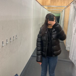 Svart pufferjacka - Säljer en snygg svart pufferjacka med dragkedja. Jackan har en normal passform och är perfekt för kyligare dagar. Den är långärmad och har en glansig yta som ger en modern look.