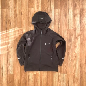 Svart vindjacka från Nike - Säljer en stilren svart vindjacka från Nike med huva och dragkedja. Jackan har långa ärmar och två praktiska fickor med dragkedjor. Perfekt för en sportig look eller en promenad i stan. Jackan är i storlek S men passar M