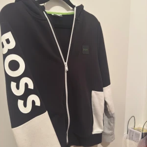 Svart Boss kofta - Snygg svart hoodie från Boss med vit dragkedja och vita detaljer på ärmarna. Stort BOSS-tryck på ena ärmen ger en cool look. Perfekt för en avslappnad stil.