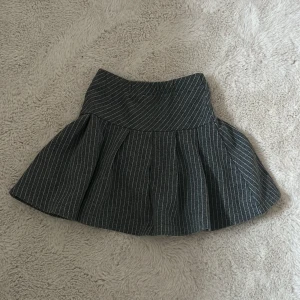 Grå kortkjol med vita ränder från ZARA - Säljer en grå barn kortkjol från ZARA med tunna vita ränder. Kjolen har en markerad midja och utställd form som ger en snygg siluett. Perfekt för dig som gillar en klassisk men trendig look. 