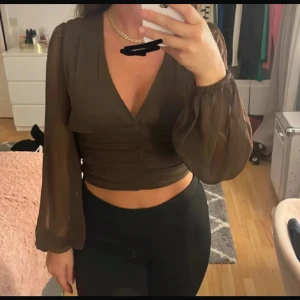 Brun croppad blus med transparenta ärmar  - Säljer en brun croppad blus med djup v-ringning och långa, transparenta ärmar från BikBok i storlek XS. Toppen har en åtsittande passform i midjan och är perfekt för att skapa en trendig look.