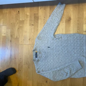 Ljusgrå kabelstickad tröja från Polo Ralph Lauren - Säljer en klassisk ljusgrå kabelstickad tröja från Polo Ralph Lauren. Tröjan har lång ärm, rund halsringning och den ikoniska broderade loggan på bröstet. Perfekt för en stilren och tidlös look.