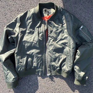 Grön bomberjacka från 157 - Säljer en klassisk grön bomberjacka från 157 med orange quiltat foder. Jackan har dragkedja framtill, ribbade muddar och en ficka med dragkedja på ärmen. Perfekt för dig som gillar streetwear och vill ha en tidlös look.