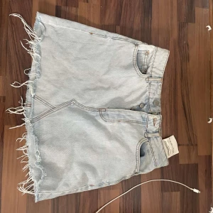 Ljusblå jeanskjol från Bershka Vintage - Ljusblå jeanskjol från Bershka Vintage med fransad nederkant och klassiska fem fickor. Kjolen har knapp- och dragkedjestängning framtill och är tillverkad i denim. Perfekt för en avslappnad stil.