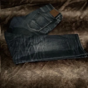 Mörkblå jeansbyxor med klassisk femficksdesign - Säljer ett par mörkblå jeansbyxor med klassisk femficksdesign och läderpatch bak i midjan. Byxorna har raka ben och tydliga slitningar för en cool look. Perfekta till vardags och passar till det mesta.