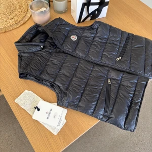 Svart dunväst från Moncler - Säljer min Moncler väst då den är för liten för mig. Den är i toppskick förutom två små lagningar vid vänster ficka. Kvitto, dustbag och NK påse följs med vid köp. Perfekt väst inför sommaren! Hör av vid funderingar👌🏻