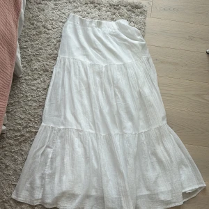 Vit långkjol i linne - Säljer en luftig och somrig vit långkjol i linne med volangdetaljer. Kjolen har en avslappnad passform och resår i midjan för extra komfort. Perfekt för varma dagar. Använd en gång. 