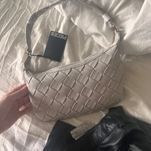 Beige axelväska från Pieces - Säljer en grå axelväska från Pieces med flätad design och justerbart axelband. Väskan har dragkedja upptill och är perfekt för att bära dina viktigaste saker. Enkel och stilren look som passar till många outfits. Pris kan diskuteras vid snabb affär 