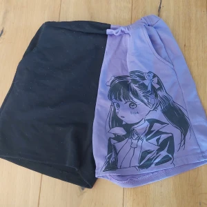 Svart och lila shorts med anime-tryck -  shorts med ena sidan i svart och andra i lila med stort anime-tryck av en tjej med rosetter i håret. Shortsen har resår. Perfekt för dig som gillar japansk stil och vill sticka ut.