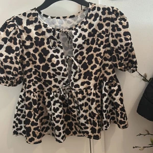Leopardmönstrad blus med knyt - Säljer en trendig blus med leopardmönster och puffiga korta ärmar. Blusen har tre dekorativa knyt framtill och en lätt utställd passform nedtill. Perfekt för dig som vill sticka ut med ett djurmönster! Oanvänd 